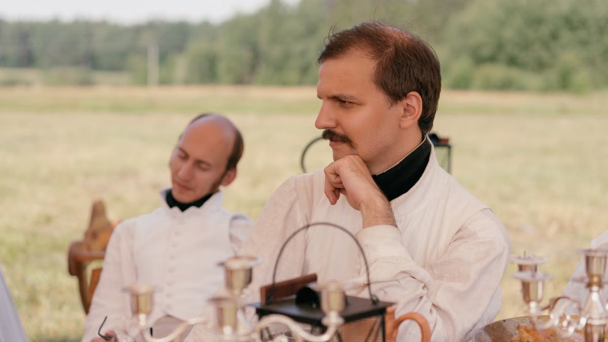 Two men in historical attire sit at a table outdoors, engaging in a reenactment.